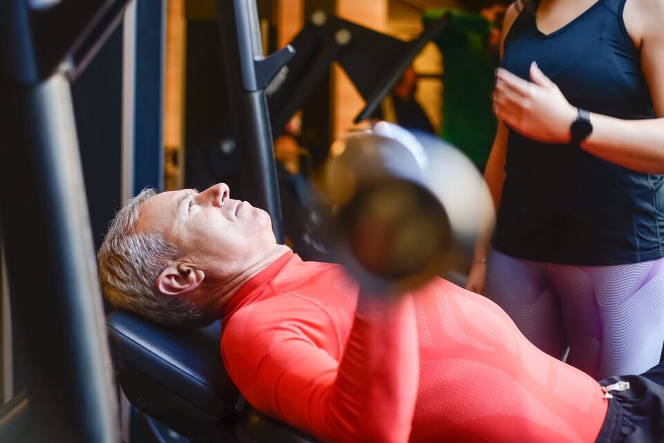 A coach supporting an older man during a bench-press exercise in a gym.