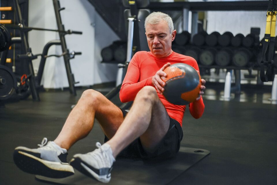 An older man performs a seated medicine-ball rotation on an exercise mat.