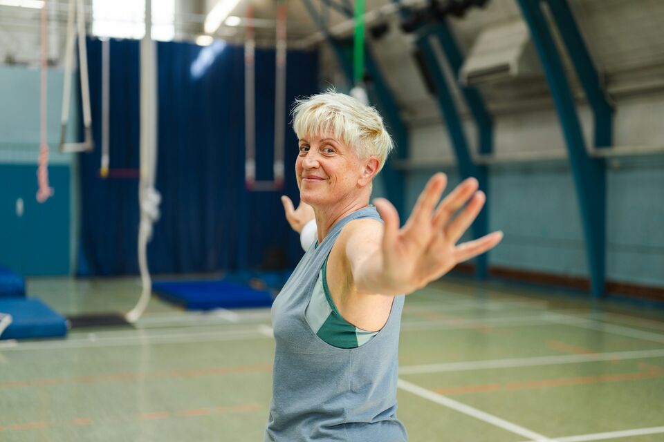 An older woman holds her arms wide during a balance exercise in a sports hall.